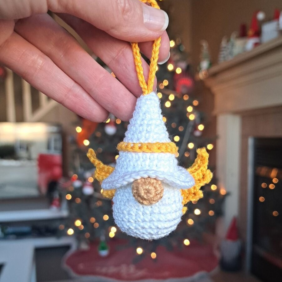 A hand holds a small crocheted Angel ornament in front of a blurred Christmas tree and fireplace. The ornament is white with yellow trim, a yellow hat, and a gold circular nose detail.