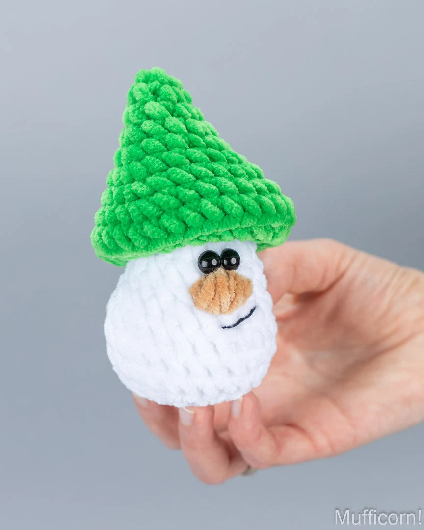 A small crocheted snowman toy with a green hat and white body, held in a person's hand against a plain gray background.