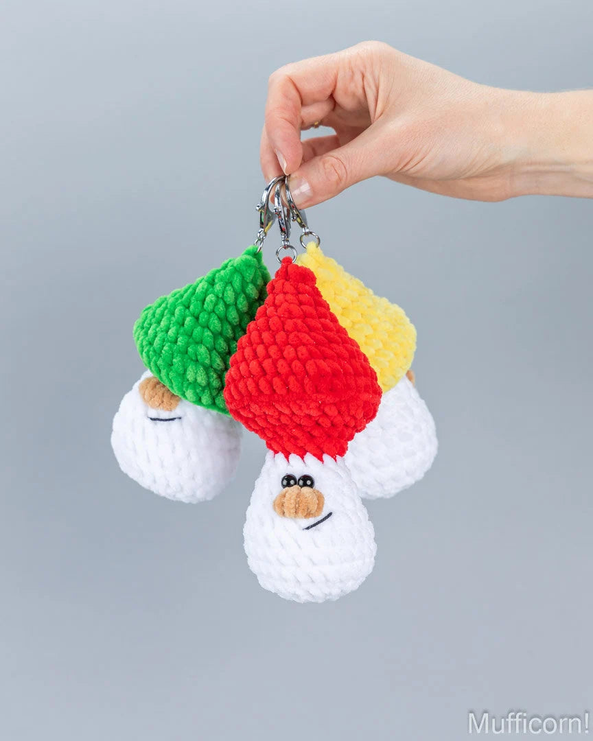A hand holding a set of three crocheted mushroom keychains with colorful red, green, and yellow hats on a plain gray background.