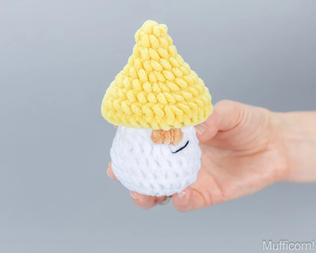 A hand holding a small crocheted gnome figure with a yellow pointed hat and white beard against a plain gray background.