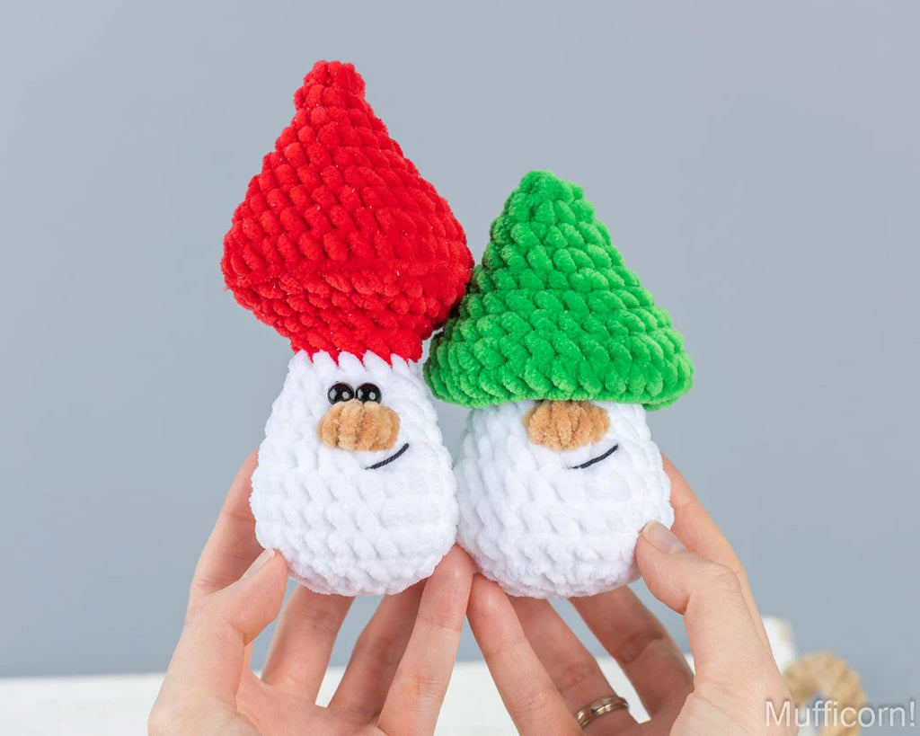 Two small crocheted elf toys being held up by hands against a soft gray background. One elf has a red hat and the other has a green hat, both with white bodies and embroidered faces.