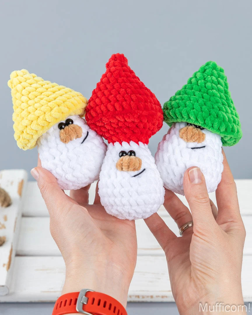 Three small crocheted gnome figures with white bodies and pointed hats in yellow, red, and green, held in two hands against a light gray background.