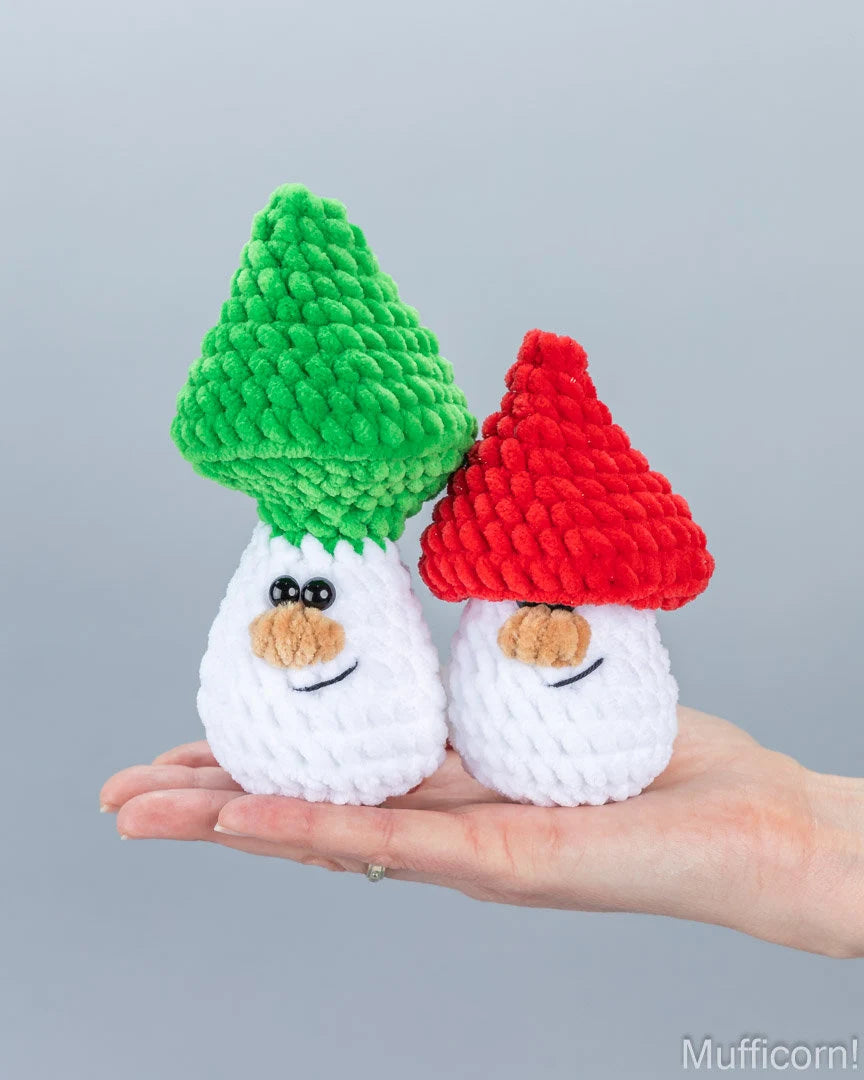 Two small crocheted gnome ornaments with white bodies, one with a green hat and one with a red hat, held in a person's hand against a plain gray background.