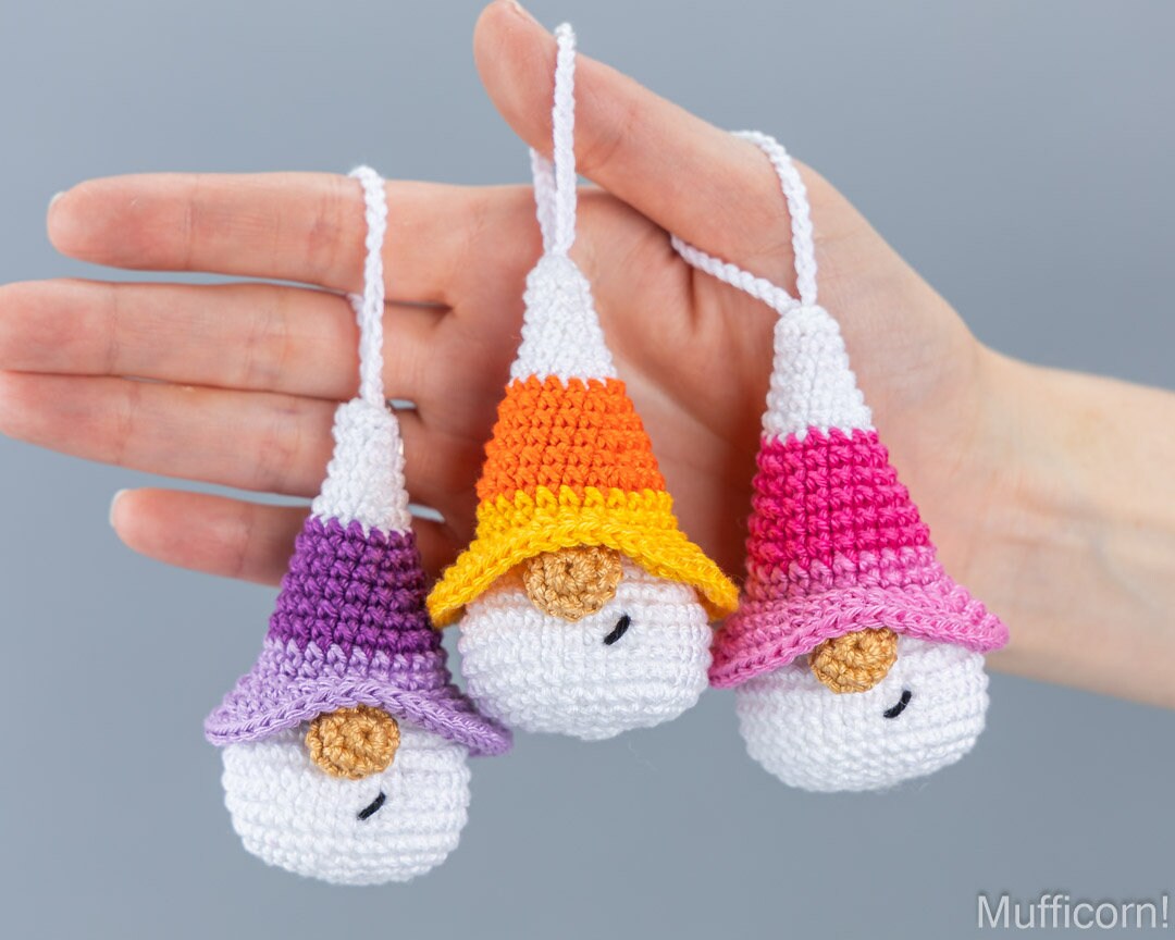 Three small crocheted gnome ornaments are held in a person's hand against a plain gray background. Each ornament has a white body, a pointed striped hat in different colorways (purple, orange/yellow, pink), and a small embroidered face.
