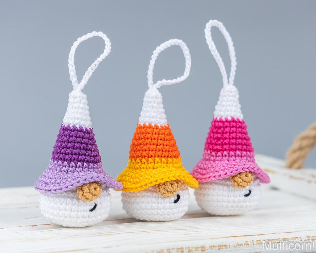 Three crocheted gnomes wearing candy corn hats in purple, orange, and pink, arranged on a white wooden surface against a soft gray background.