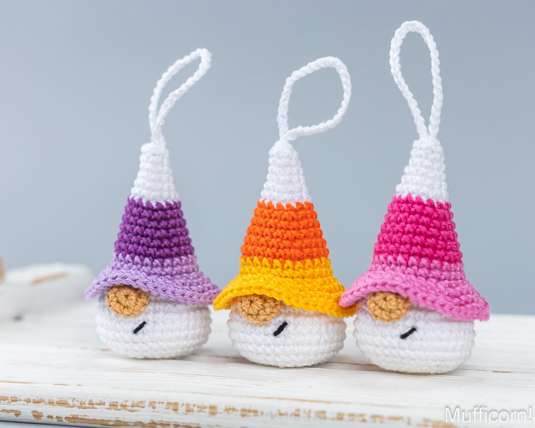 Three small crocheted candy corn ornaments with hanging loops, displayed on a white wooden surface against a soft light gray background. Each ornament has a white body, a pointed corn-hat shape in different color combinations, and embroidered eyes and noses.