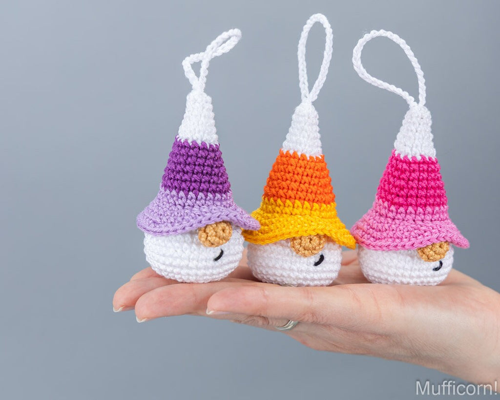 Three small crocheted muffin ornaments shaped like candy corn are displayed on a person's hand against a plain gray background. The ornaments have white bodies, pointed hats in purple, orange, and pink, and small tan faces.