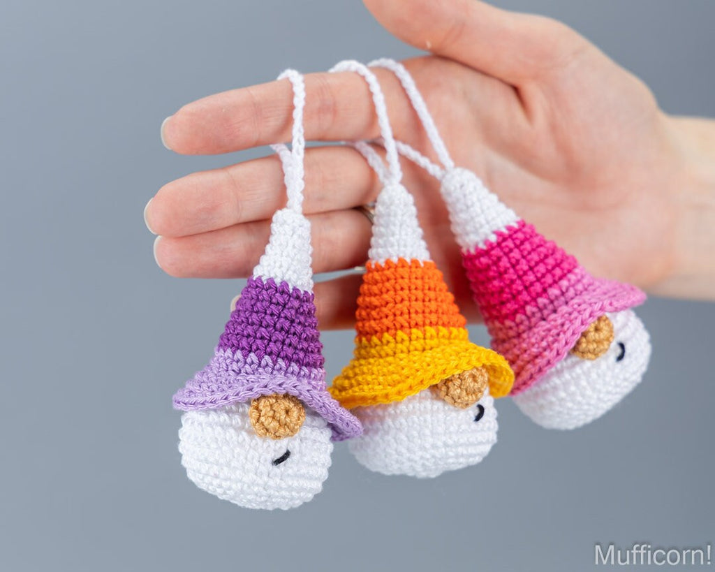Three crocheted muffin-shaped keychains or ornaments are held in a person's hand against a plain gray background. Each has a white body, a pointed hat in different colors (purple, orange/yellow, pink), and a small brown crumb detail.