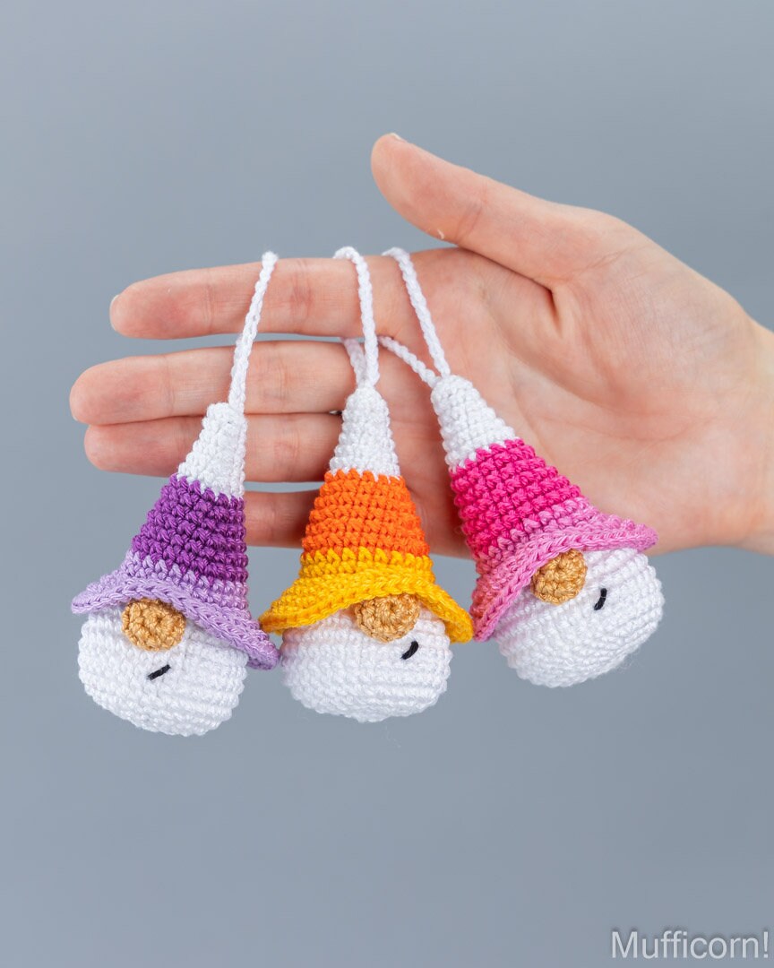 Three crocheted gnome keychains in purple, orange, and pink are held in a person's hand against a solid gray background.