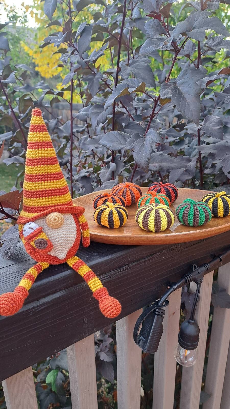 A crocheted gnome doll with a striped orange and yellow hat sits on a wooden deck railing next to a wooden tray holding several small crocheted pumpkins, with garden foliage in the background.