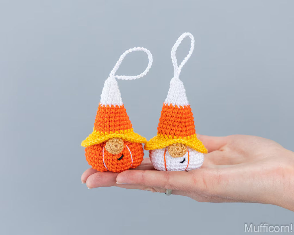 Two small crocheted candy corn gnomes with white hanging loops are held on a person's hand against a plain light gray background. The product color identity is orange, white, and yellow, mimicking the candy corn design.