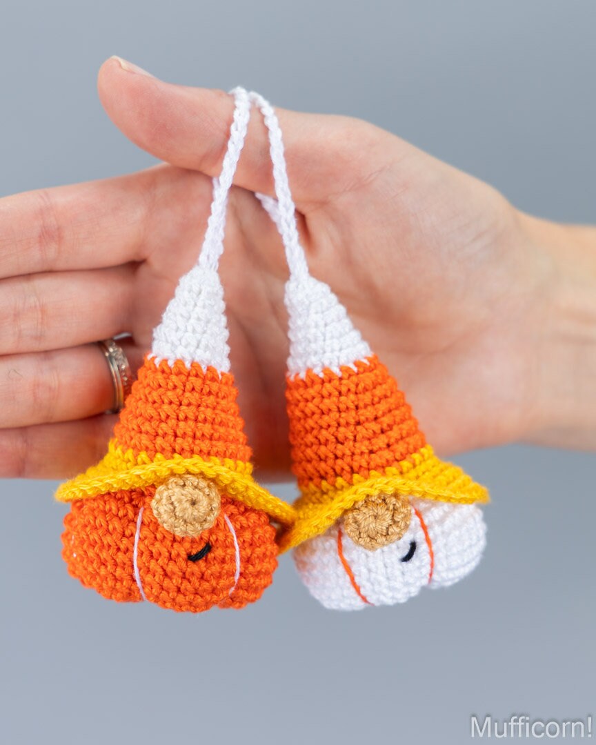 A pair of handmade crocheted candy corn gnomes in orange, white, and yellow, held in a person's hand against a solid gray background.