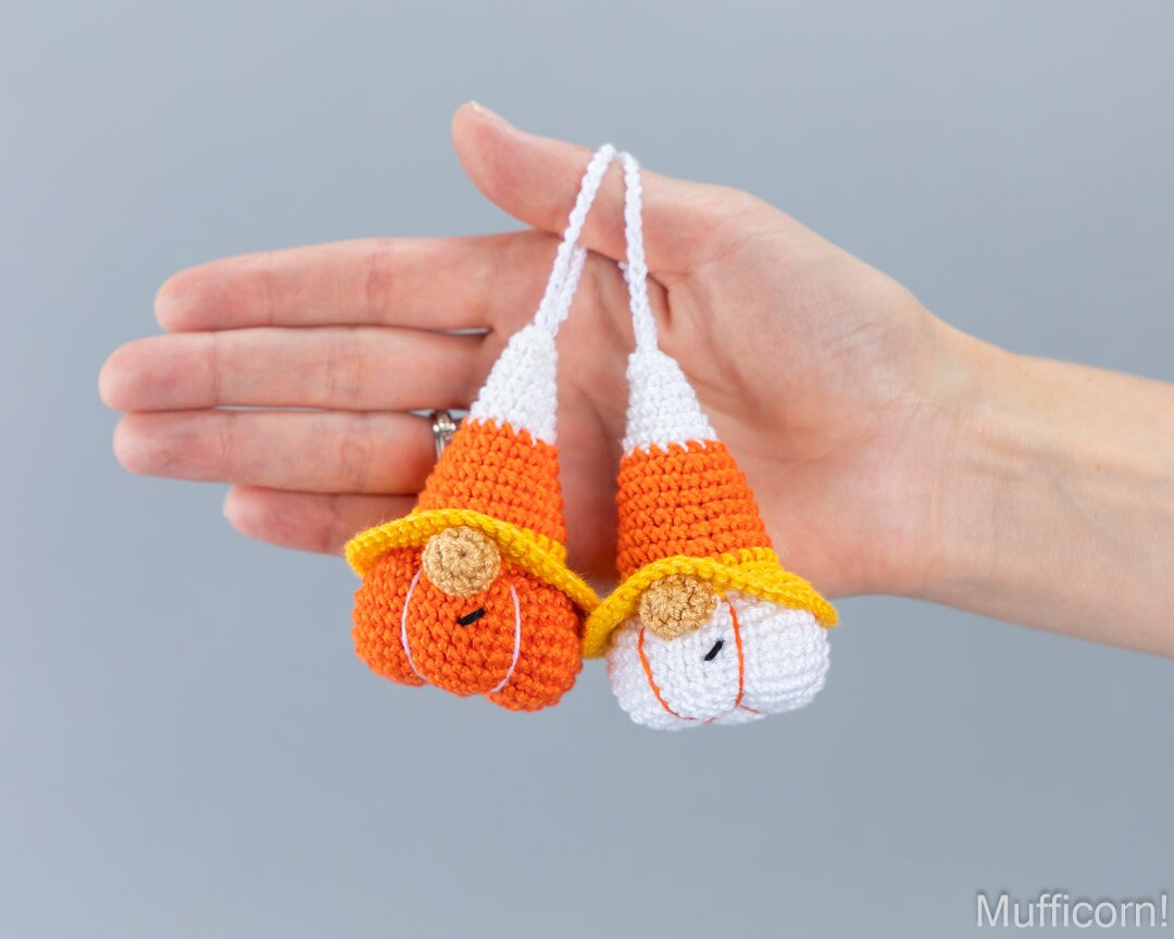 A hand holds two small crocheted candy corn gnomes shaped like pumpkins and candy corn, with orange, white, and yellow colors, against a solid gray background.