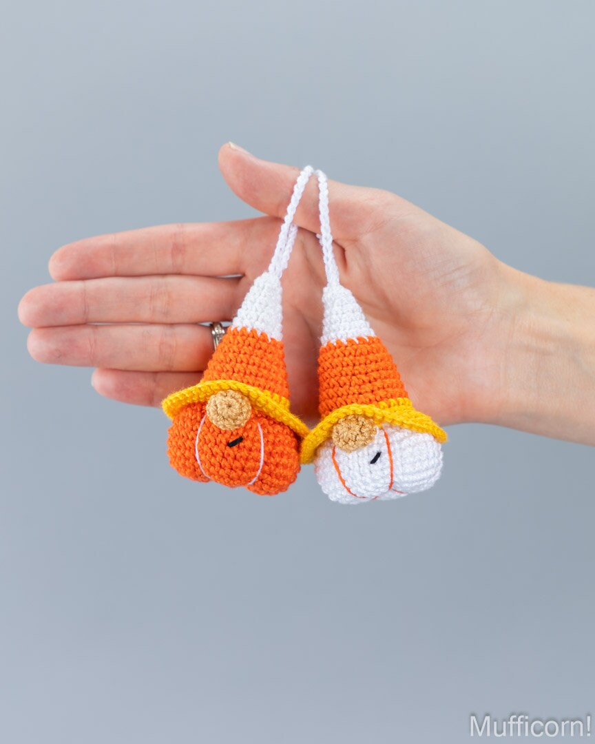 A hand holding two small crocheted candy corn gnomes ornaments, one orange and white with a pumpkin face, and one orange and white with a candy corn face, against a plain gray background.