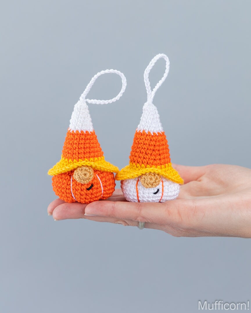 Two small crocheted candy corn gnomes held in a person's hand against a plain light gray background. One gnome is orange and white with a yellow hat, and the other is white and orange with a yellow hat.