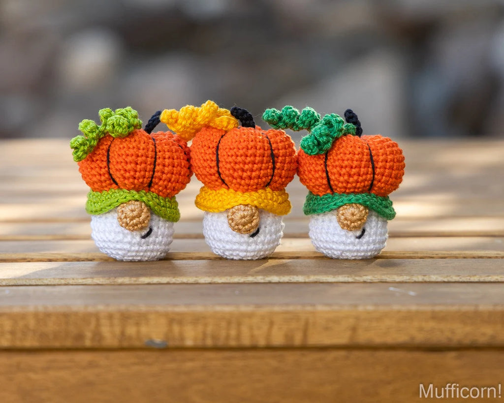 Three small crocheted pumpkin gnome figures sitting side by side on a wooden surface, with a blurred outdoor background. The word 'Mufficorn!' appears in the bottom right corner.