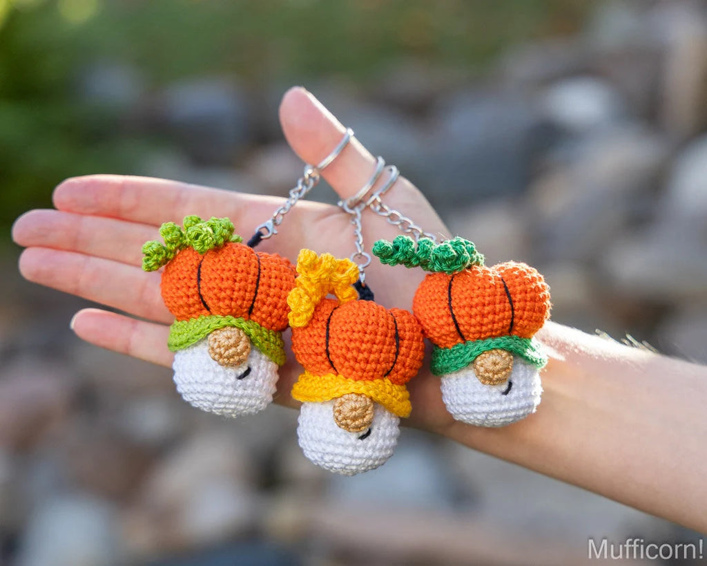Three small crocheted pumpkin keychains with smiling faces are held in a person's hand outdoors. Each pumpkin is orange with green and yellow tops, and the keychains are attached to silver metal rings.