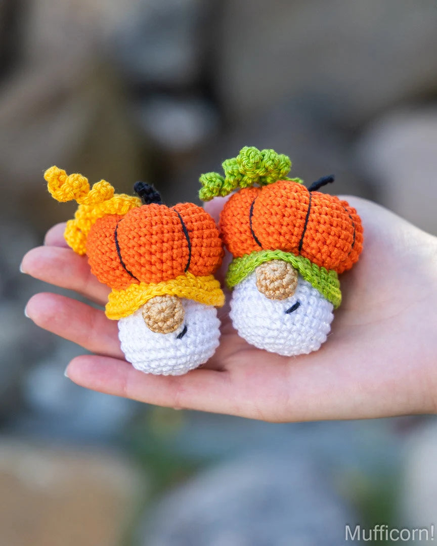 Two small crocheted pumpkin gnomes held in a person's hand against a blurred outdoor background. Each gnome has an orange crocheted pumpkin body with black stitched lines, a white face, a yellow hat, and one has a green hat and the other has a yellow hat.