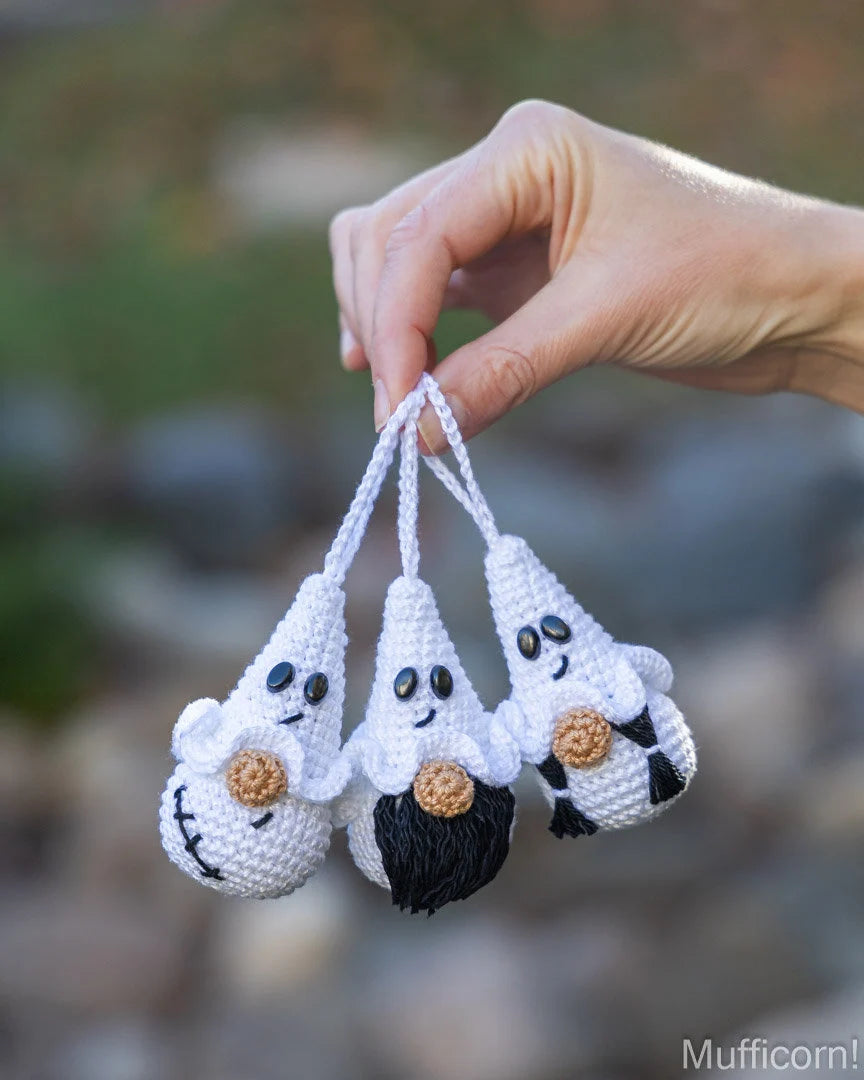 Three small white crocheted ghost ornaments with black and brown details are being held by a person's hand against a blurred outdoor background.