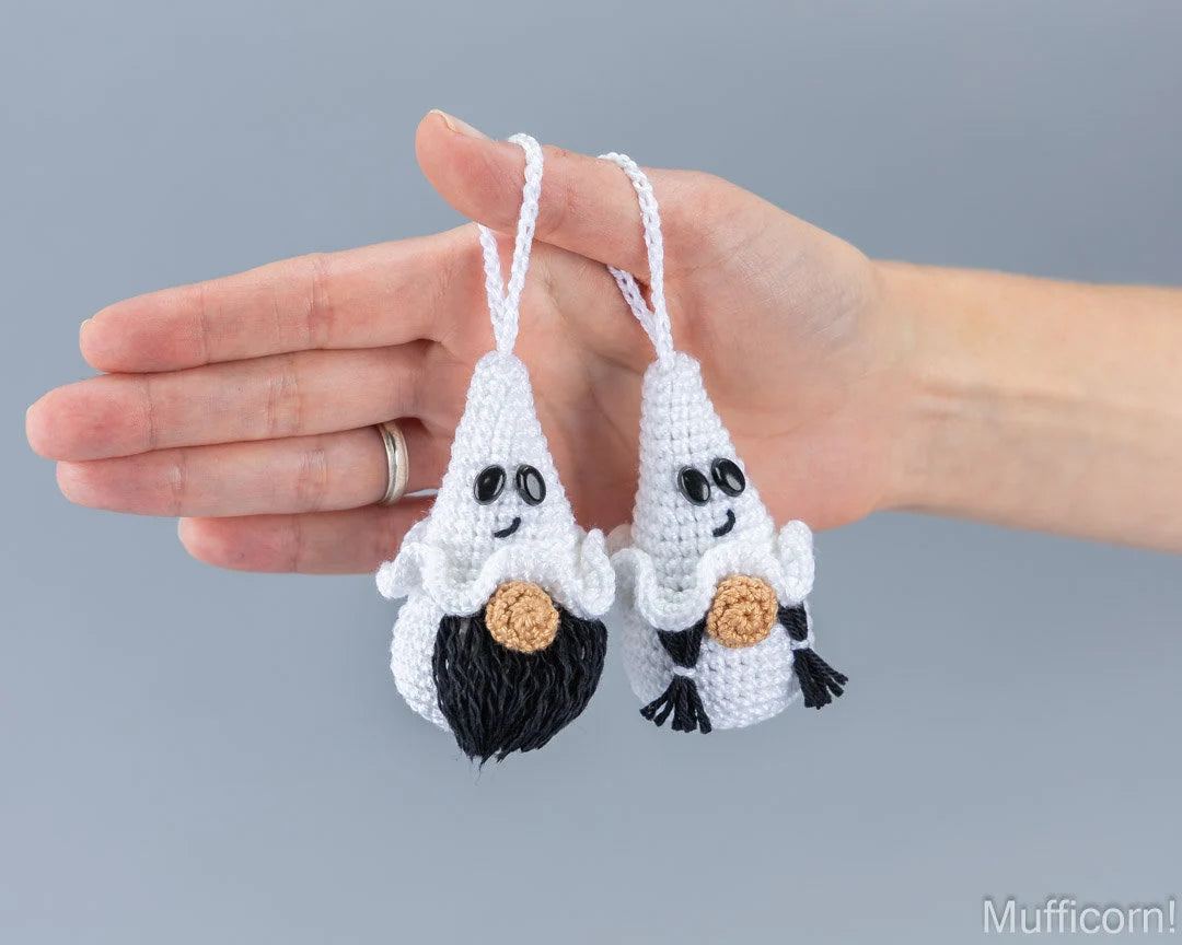 Two small crocheted ghost ornaments being held in a person's hand against a plain gray background. The ornaments are white with black facial features, black hair, and tan circular details on their bellies.