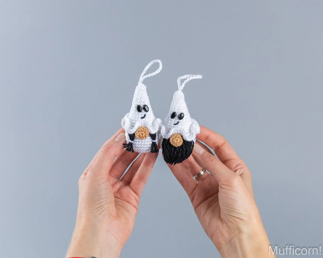 Two small crocheted ghost ornaments held up by two hands against a plain gray background. 