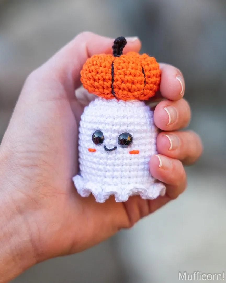 A hand holding a small crocheted toy featuring a white ghost body with black bead eyes and a bright orange pumpkin on its head, photographed outdoors against a softly blurred background.