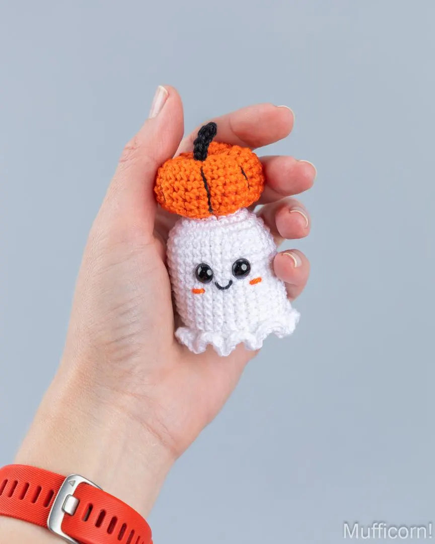 A hand holds a crocheted Halloween-themed amigurumi toy featuring a white ghost with a bright orange pumpkin on its head, set against a solid light gray background.