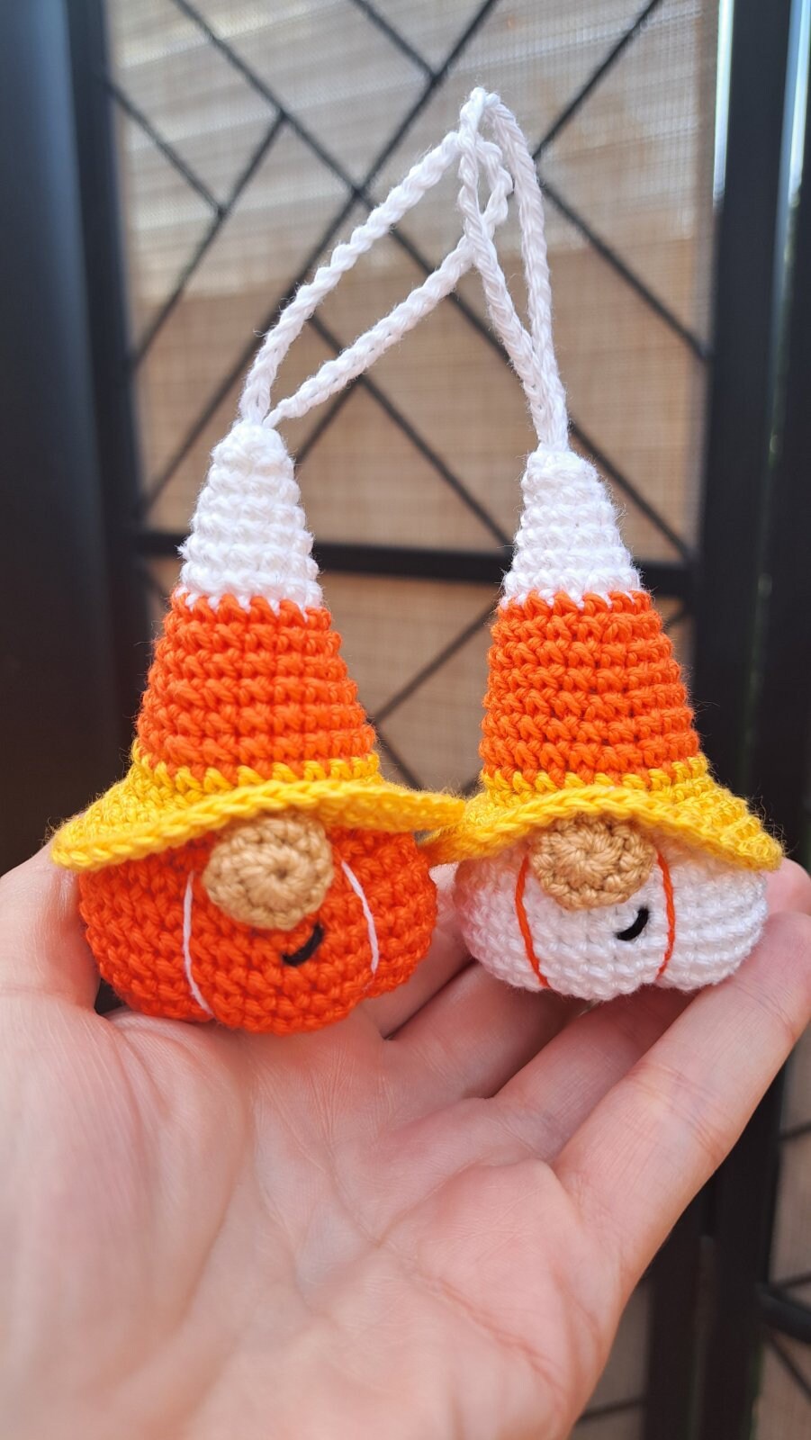 Two small crocheted candy corn gnomes held in a person's hand. Each gnome has a white and orange cone body with a yellow brim and a round tan face, and white hanging loops at the top.