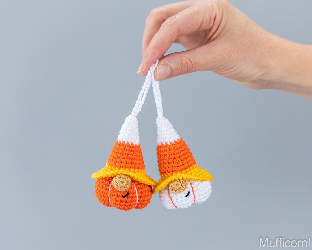 A hand holds two small crocheted candy corn gnomes shaped like candy corn, one orange and white with a yellow hat and one white and orange with a yellow hat, against a plain gray background.
