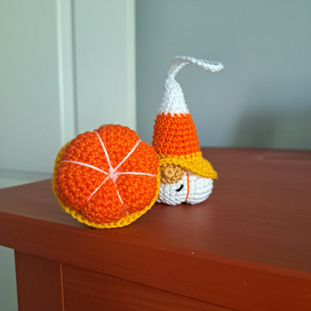A handmade crocheted amigurumi ornament shaped like a candy corn gnomem and a matching orange pumpkin ball, displayed on a wooden table against a gray wall.