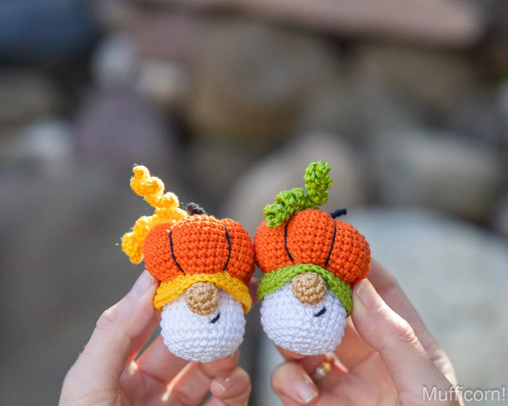 Two small crocheted pumpkin-and-gnome amigurumi toys are held in two hands against a blurred outdoor stone background. One toy has an orange pumpkin body with yellow-orange stem and hat, and a white face.
