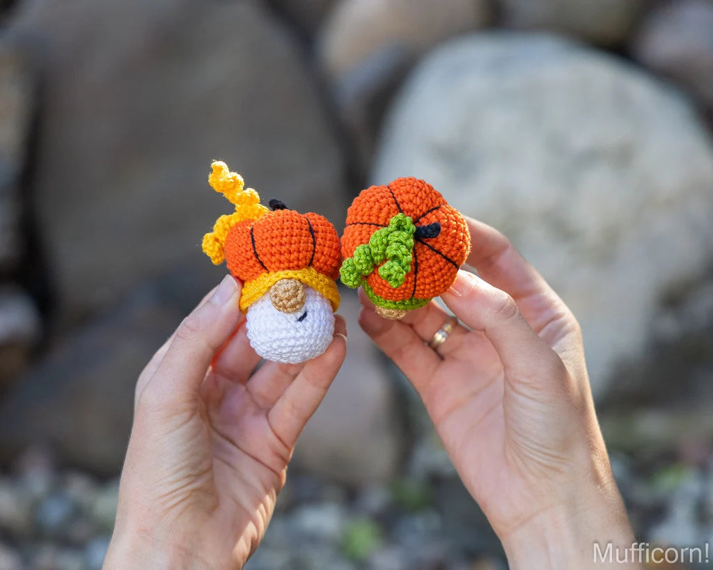Two hands holding two small crocheted pumpkin characters outdoors with a blurred rocky background. One pumpkin is orange with yellow stems and a white face, and the other is orange with green leaves and a yellow stem.
