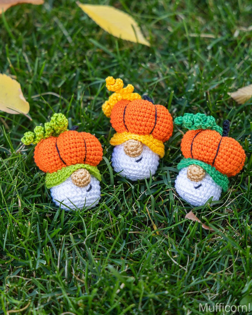 Three small crocheted pumpkin gnomes with orange bodies, white hats, and colorful leafy tops are placed on green grass with scattered yellow leaves. A 'Mufficom!' watermark appears in the bottom right corner.
