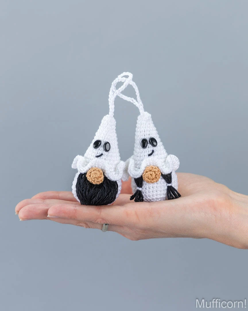 Two small crocheted ghost-shaped ornaments are held in a person's hand against a plain gray background. 
