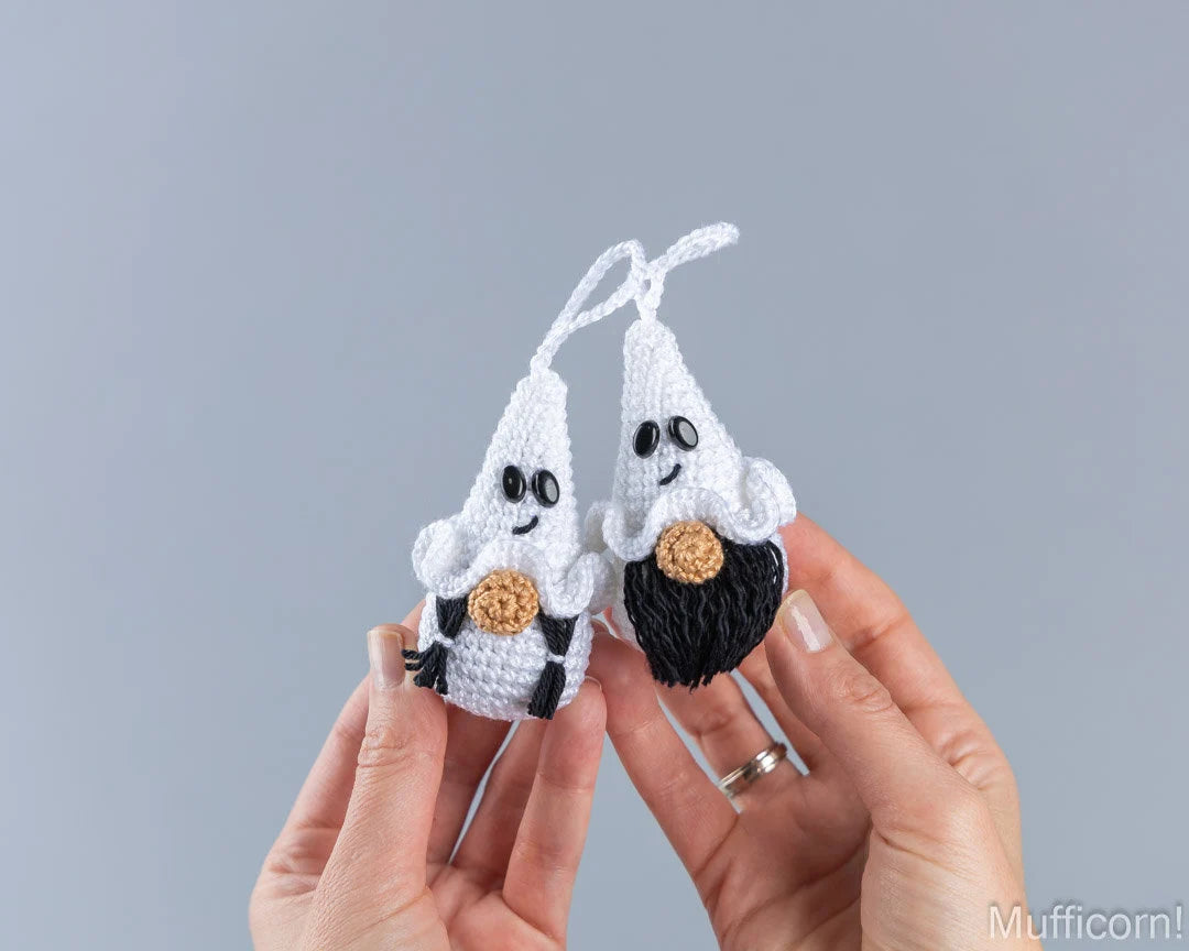 Two small crocheted ghost halloweenornaments being held in two hands against a plain gray background. 