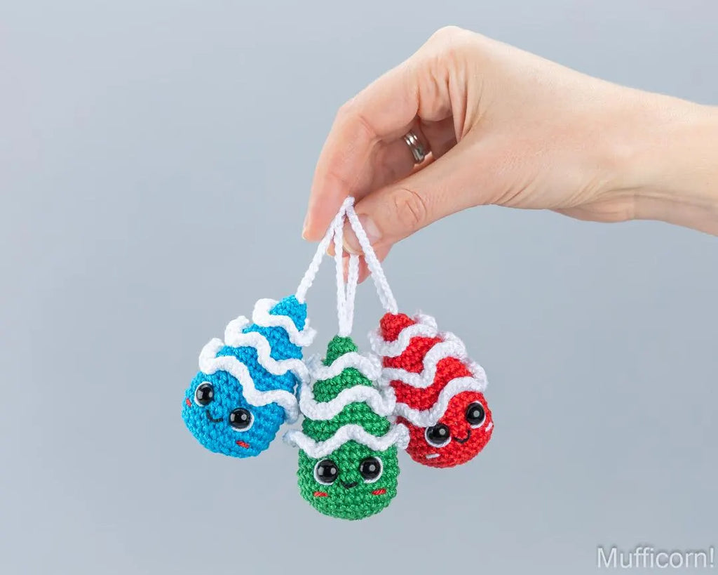 A hand holds three small crocheted character ornaments against a plain light gray background. The ornaments have wavy stripes in blue, green, and red with white accents and cartoon eyes.
