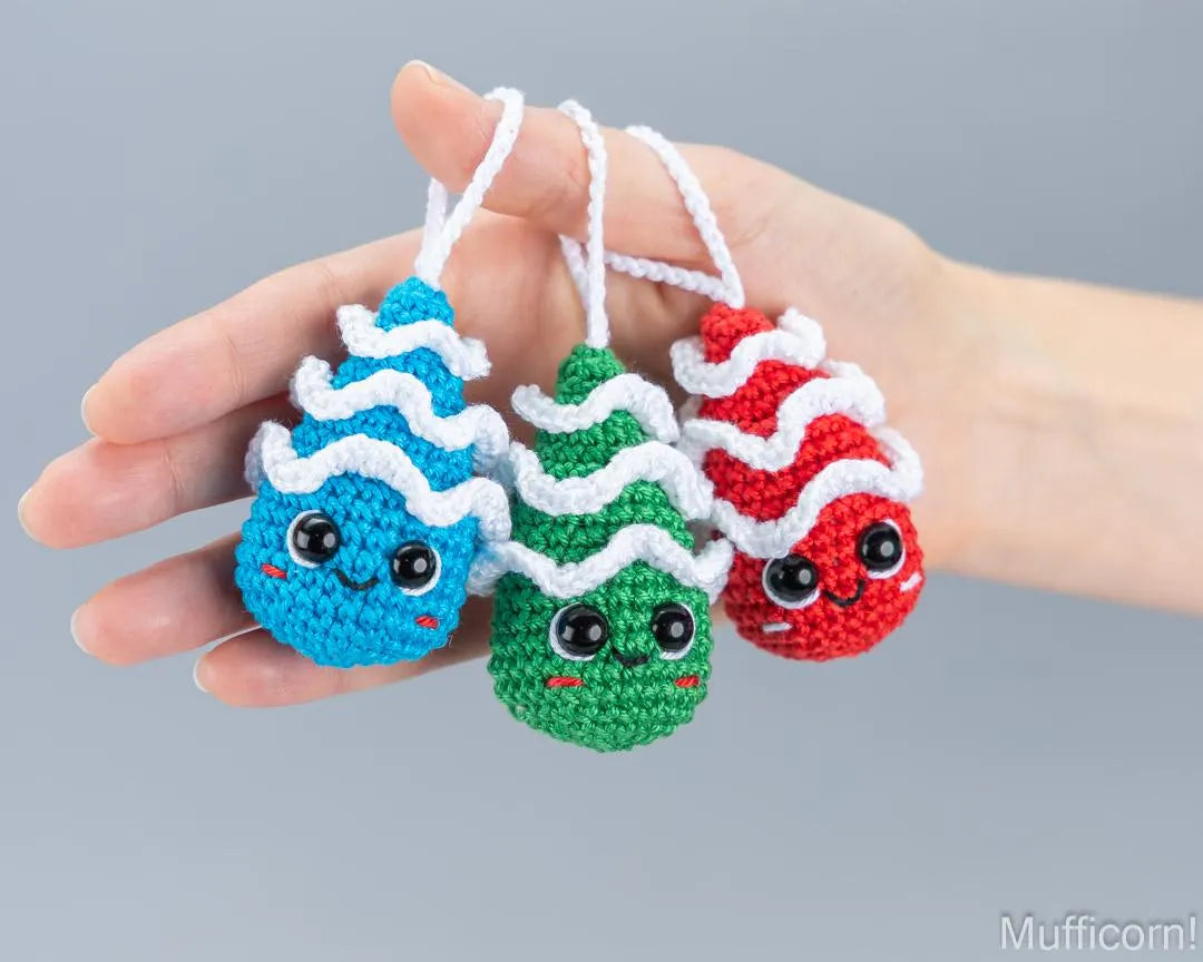 Three small crocheted character ornaments in blue, green, and red are held in a person's hand against a plain gray background. Each ornament has a white wavy yarn top and a white hanging loop.