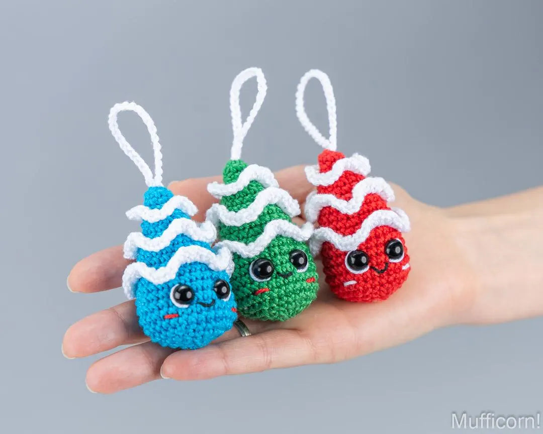 Three small crocheted chtistmas tree ornaments  in blue, green, and red, each with a white wavy top and hanging loop, held in a person's hand against a plain gray background.