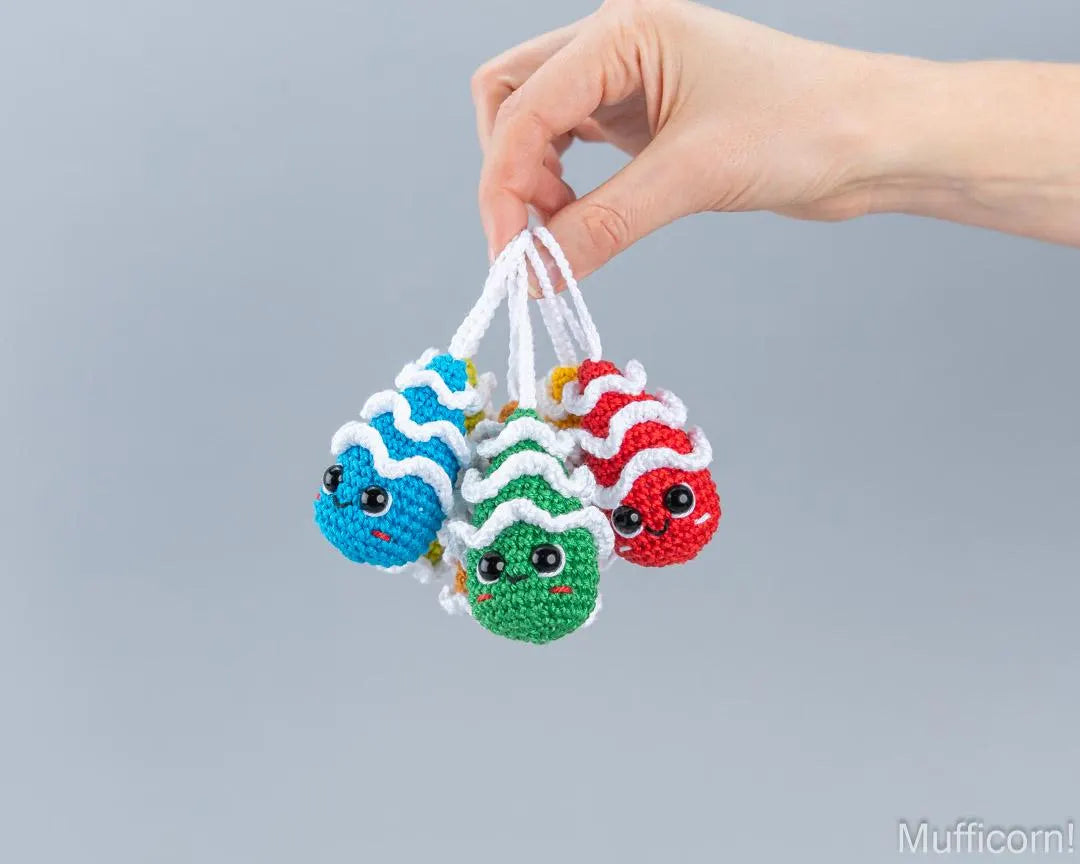 A hand holding four small crocheted fish charms in blue, green, and red, each with white wavy stripes and black eyes, against a plain gray background.