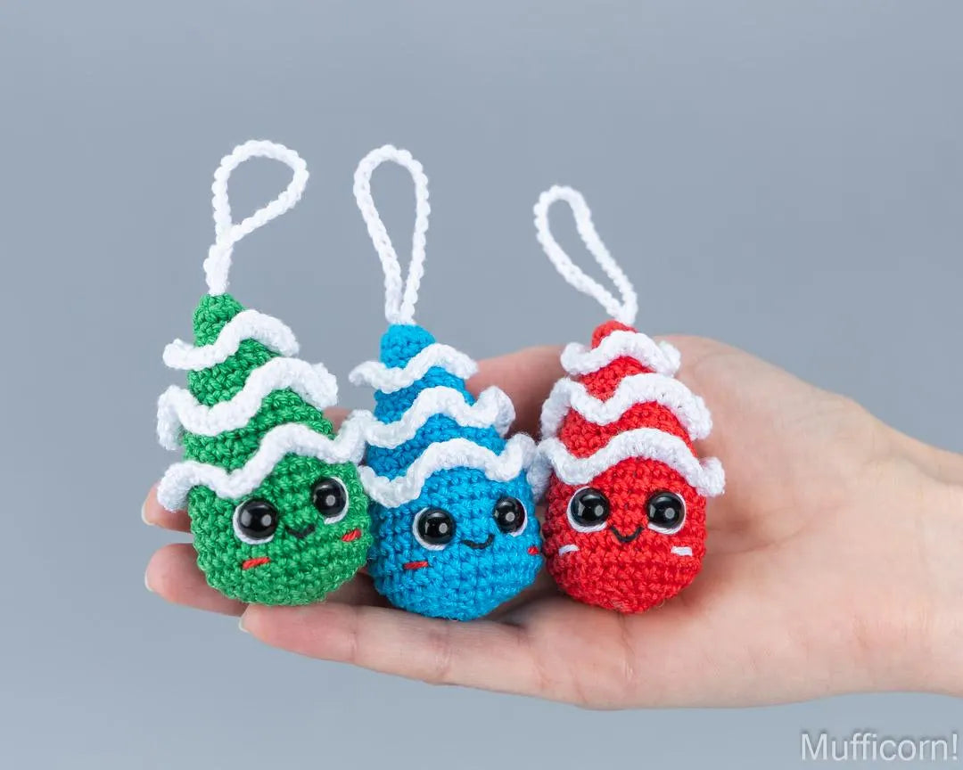 Three small crocheted Christmas tree ornaments in green, blue, and red, each with a white wavy trim and a white hanging loop, held in a person's hand against a plain gray background.