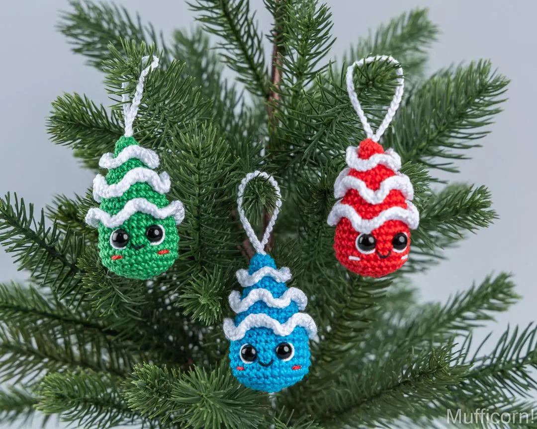 Three cute, hand-crocheted Christmas ornaments shaped like pine trees hang on a green Christmas tree branch. The ornaments are in green, blue, and red, each with white wavy details representing snow or frost, and a small embroidered face.