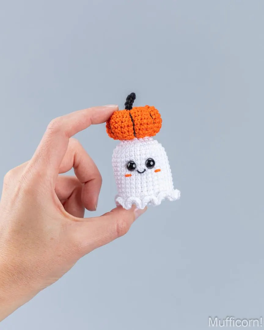 A hand holds a small crocheted ghost and pumpkin charm against a plain light gray background. The charm features a white ghost body with embroidered eyes and smile, topped with an orange pumpkin and a short black stem.