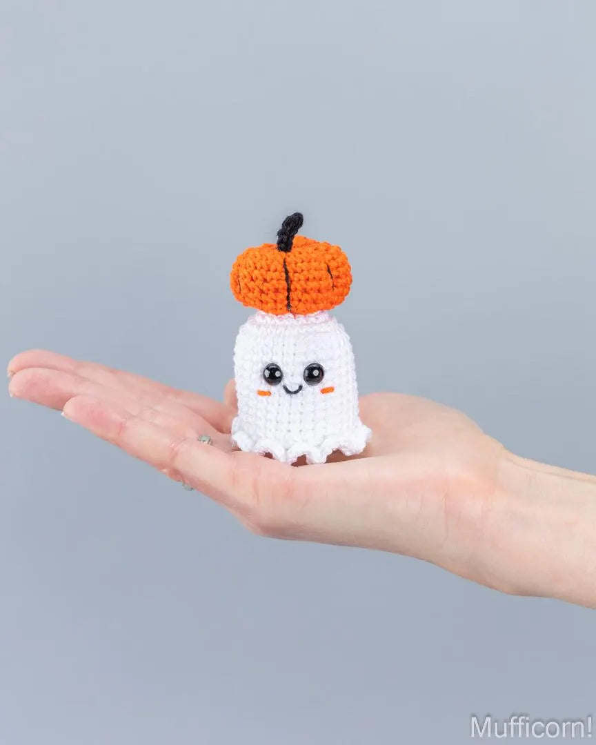 A small crocheted Halloween ghost and pumpkin figure is held in a person's open hand against a plain light gray background.
