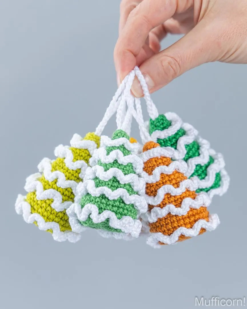 Three small crocheted christmas tree ornaments in yellow, green, and orange with white wavy trim, held by a hand against a light gray background.