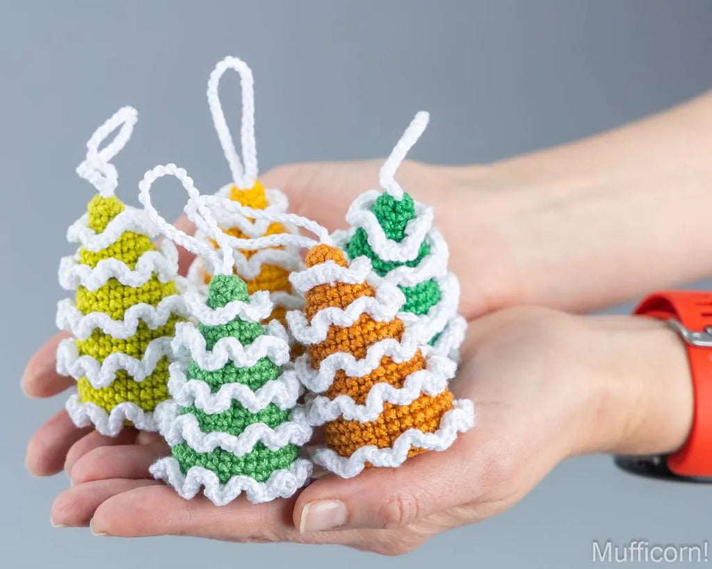 Three small crocheted Christmas tree ornaments in yellow, green, and orange with white ruffled trim are held in two hands against a plain gray background.