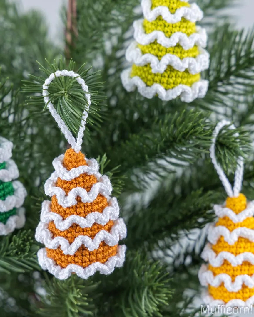 Handmade crocheted Christmas tree ornaments hanging on a green pine tree. The ornaments are shaped like pinecones with wavy striped patterns in orange, yellow, and white, with a white hanging loop.