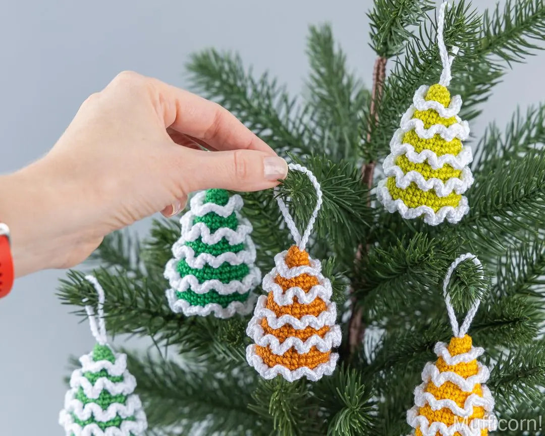 A hand is hanging a crocheted Christmas tree ornament on a green artificial tree. Several other colorful ornaments in green, yellow, and orange are already hanging on the tree against a light gray background.