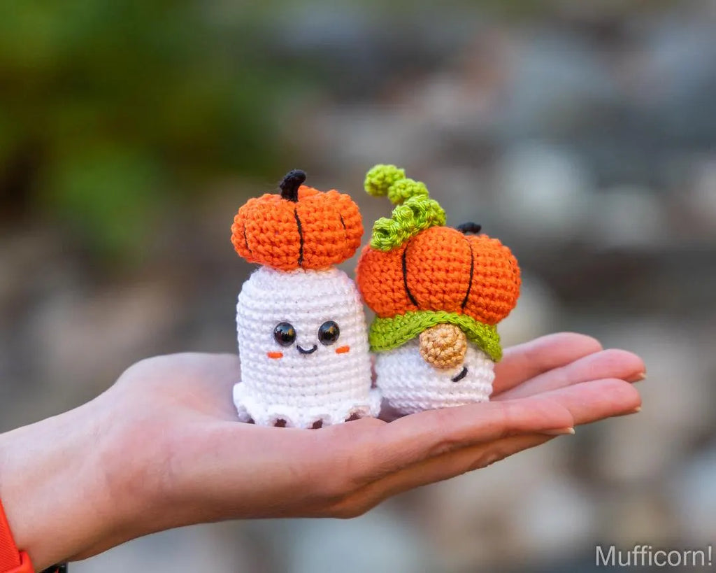Two small crocheted Halloween characters, a white ghost with an orange pumpkin and a white goblin with an orange pumpkin and green hat, held in a person's hand against a blurred outdoor background.