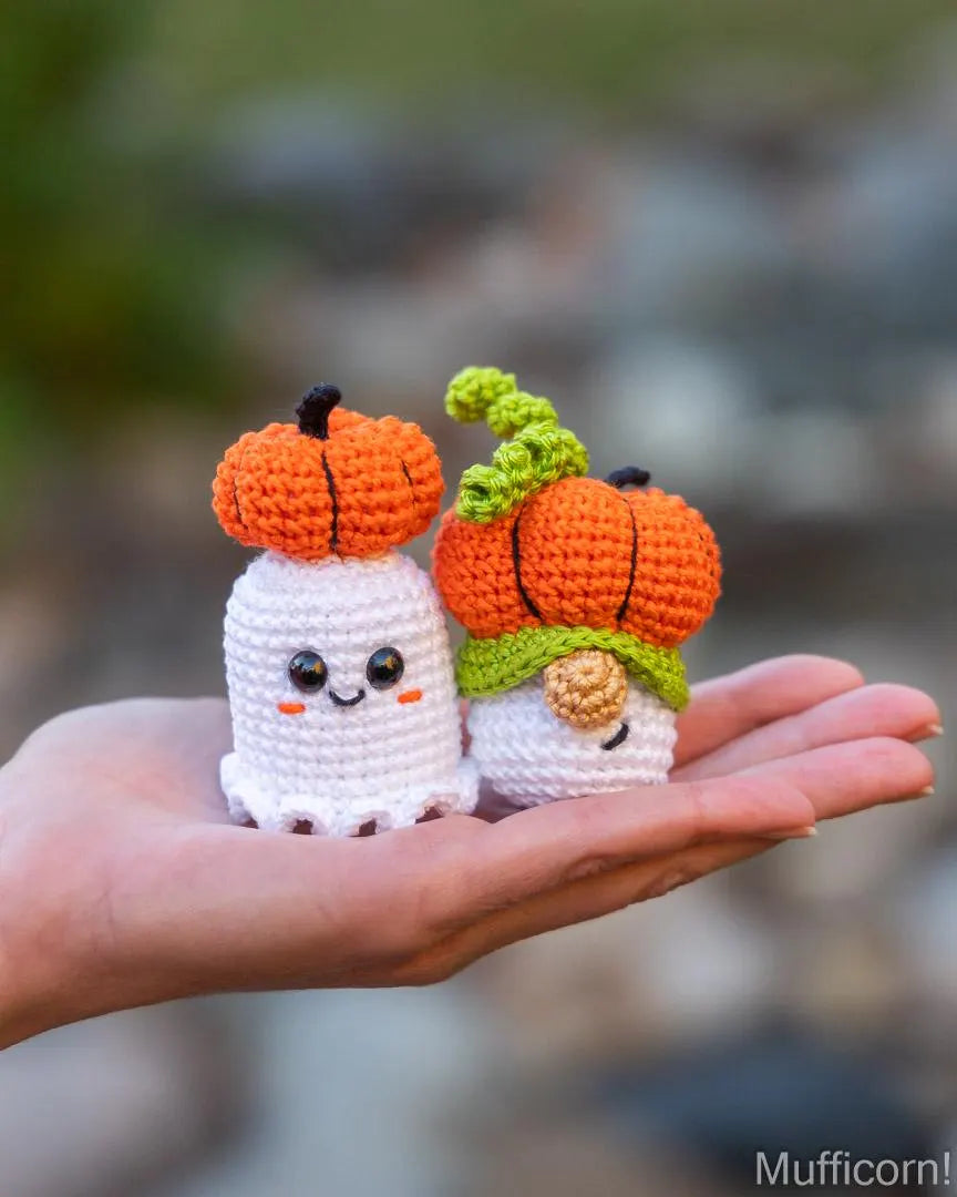 Two small crocheted Halloween characters, one white ghost with an orange pumpkin and one white gnome with an orange pumpkin and green stem, held in a person's hand against a blurred outdoor background.