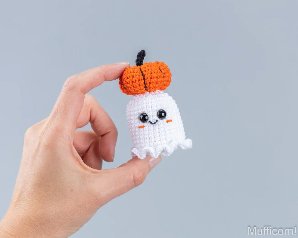 A hand holds a small crocheted Halloween ghost toy with an orange pumpkin on its head against a solid light gray background.